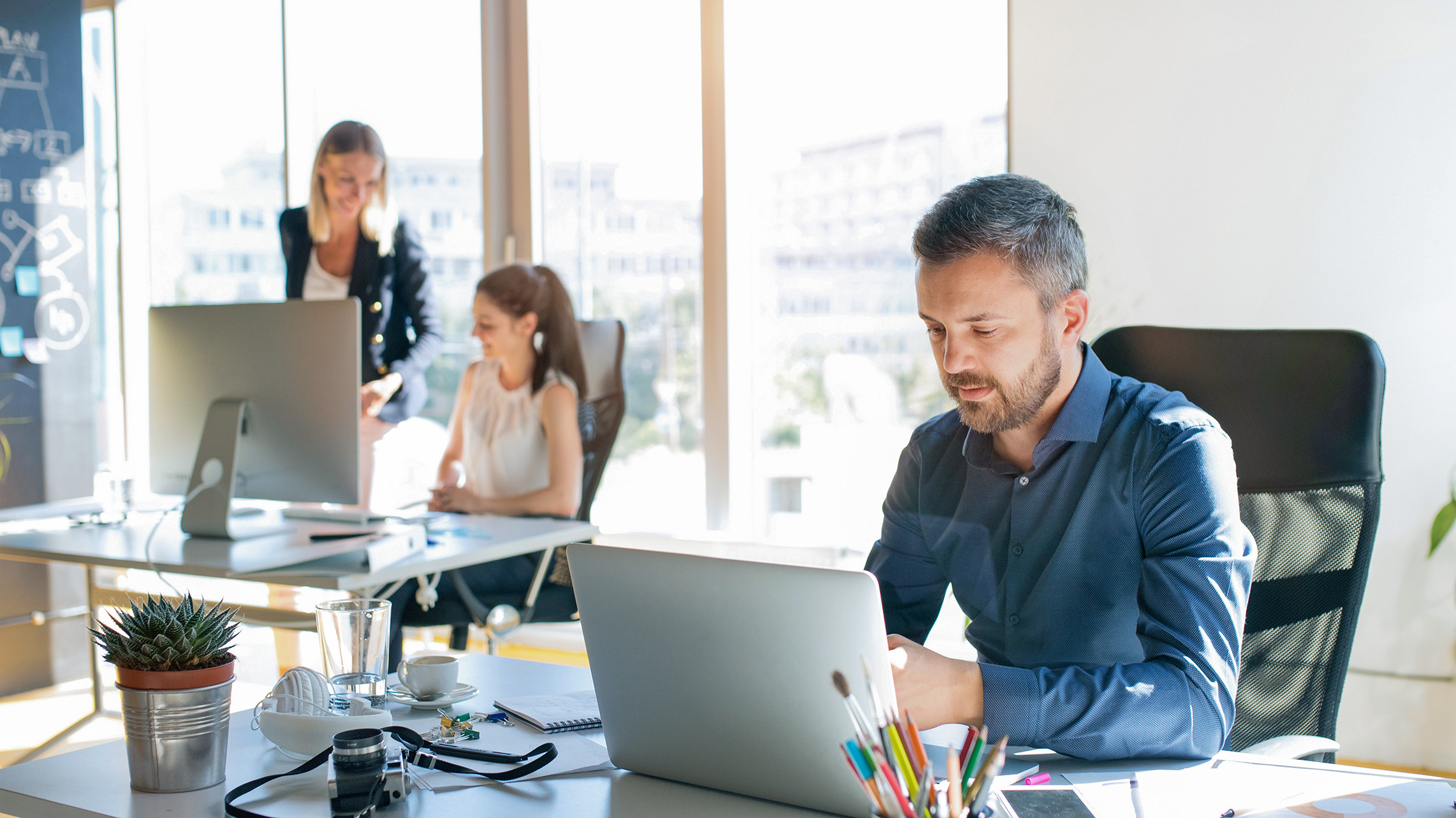 man working on a laptop in an office. team colleagues in the background. 