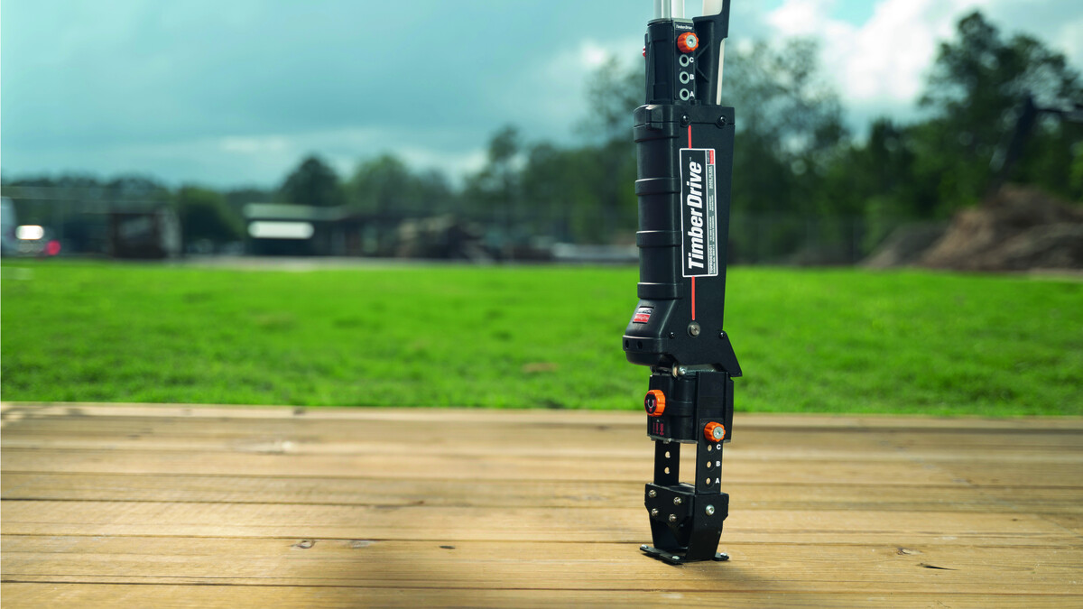 Timber-Drive main image Der Timber Drive auf einer Holzfläche, ein Tisch oder eine Werkbank. Das Gerät steht eher rechts auf dem montierten flachen Anschlag und ist aufrecht, sodass nach oben nur das Laderohr und die Schrauber-Aufnahme sichtbar sind. Nach links hin ist der Tisch leer. Hintergrund, leicht verschwommen: eine grüne Wiese, Bäume, Büsche und Gebäude und darüber der blaue, leicht bewölkte Himmel.