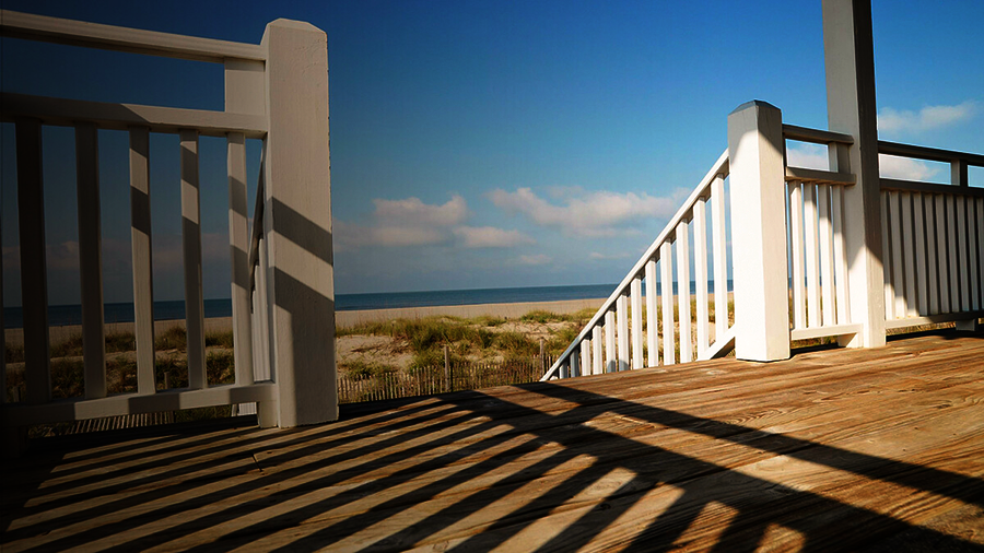 eine hölzerne Terrasse mit weißem Geländer und einer Treppe, die nach unten führt. Im Hintergrund ein Staketenzaun, dann ein spärlich bewachsener Sandstrand, dann das blaue Meer mit dem ebenfalls blauen Himmel darüber.
