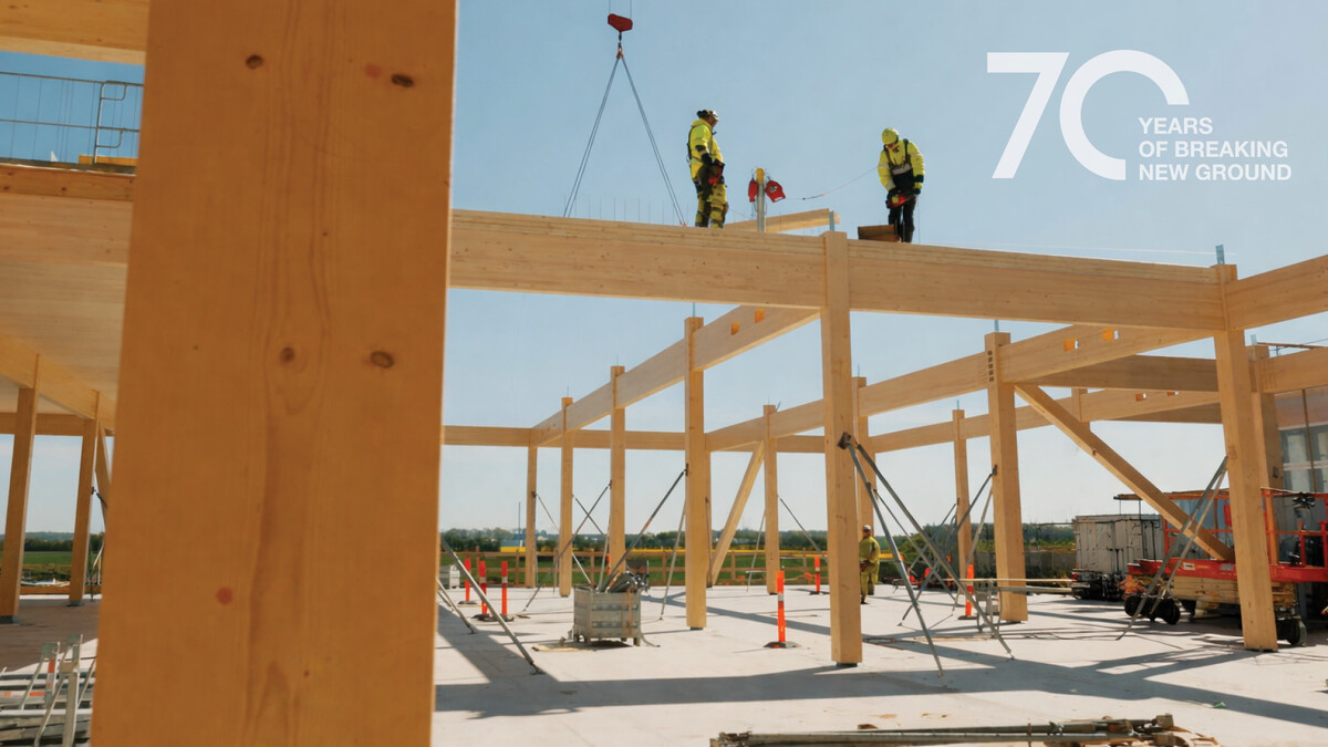 Eine Baustelle, bestehend auf einem hölzernen Gerüst. Zwei Männer in gelben Warnwesten und Sturzhelmen sind auf einem der Balken unterwegs. Der Boden besteht aus gegossenem Beton. Im rechten oberen Bildrand die Aufschrift "70 YEARS OF BREAKING NEW GROUND".