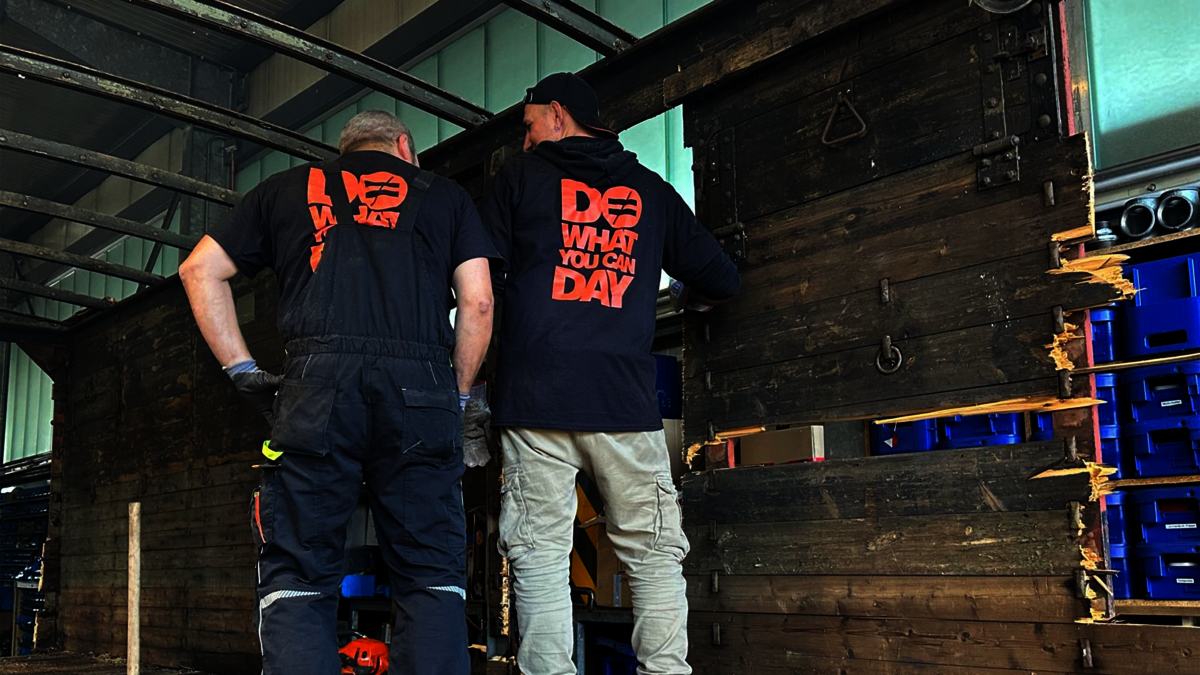 Zwei Männer stehen mit dem Rücken zur Kamera. Sie stehen in einem Waggon aus Holz.