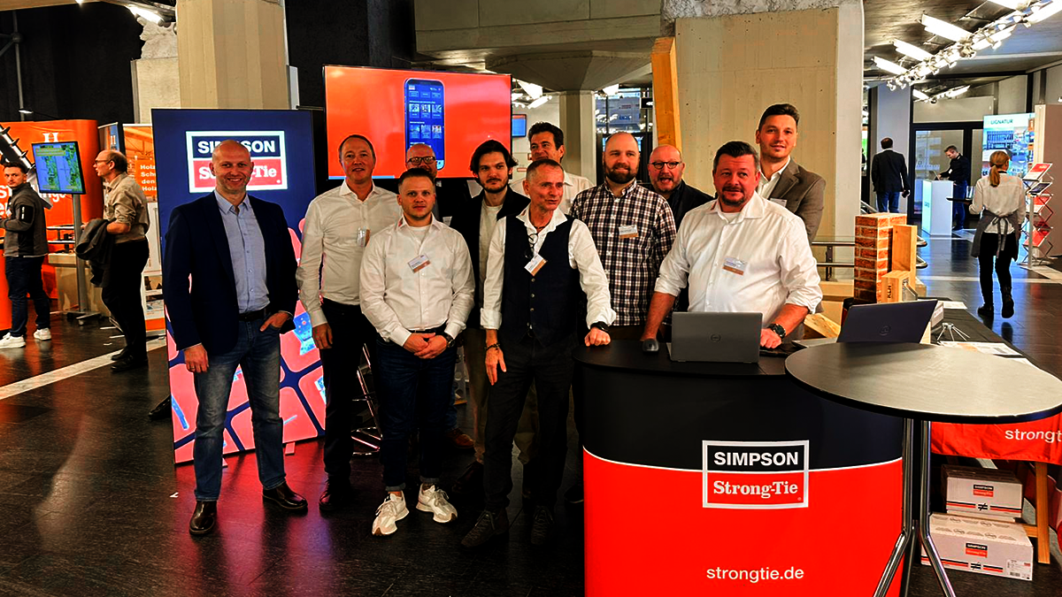 Ein Messestand mit einer rot-schwarzen Theke, einem Stehtisch, einem beleuchteten Display und einem Monitor auf einer hohen Stele. Eine zehnköpfige Mannschaft ist vor dem Stand aufgebaut und lächelt in die Kamera. Alle sind unterschiedlich gekleidet, haben aber Namensschilder am Hemd.