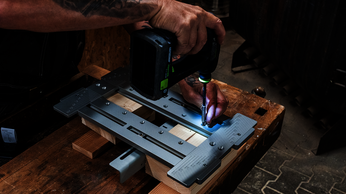 Die Fräs- und Montageschablone MIMO315 von Simpson Strong-Tie in einer Holzbauwerkstatt während der Montage eines Verbinders. Links ist eine Person zu sehen, die einen Akkuschrauber in der Hand hält und damit die Schablone auf einigen auf der Werkbank bereitliegenden Holzbauelementen fixiert.