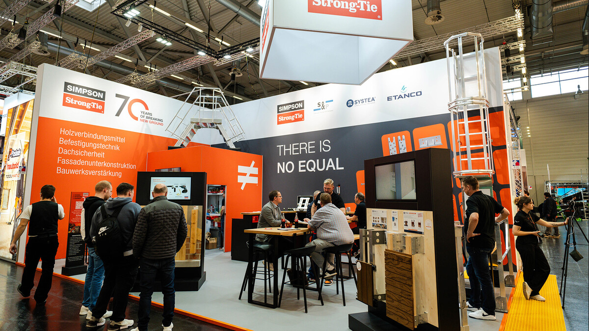 Ein Foto von einem Messestand auf der Dach+Holz 2026. Zu sehen ist der STand der Firma Simpson Strong-Tie in kräftigem Orange. Auf dem Stand befinden sich mehrere Aufsteller mit Produktmustern und Bildschirmen, außerdem gehören eine große Sicherheitsleiter und eine A-förmige Treppe aus Metall zum Stand. Viele Menschen sind zu sehen, sowohl Besucher als auch Standpersonal.
