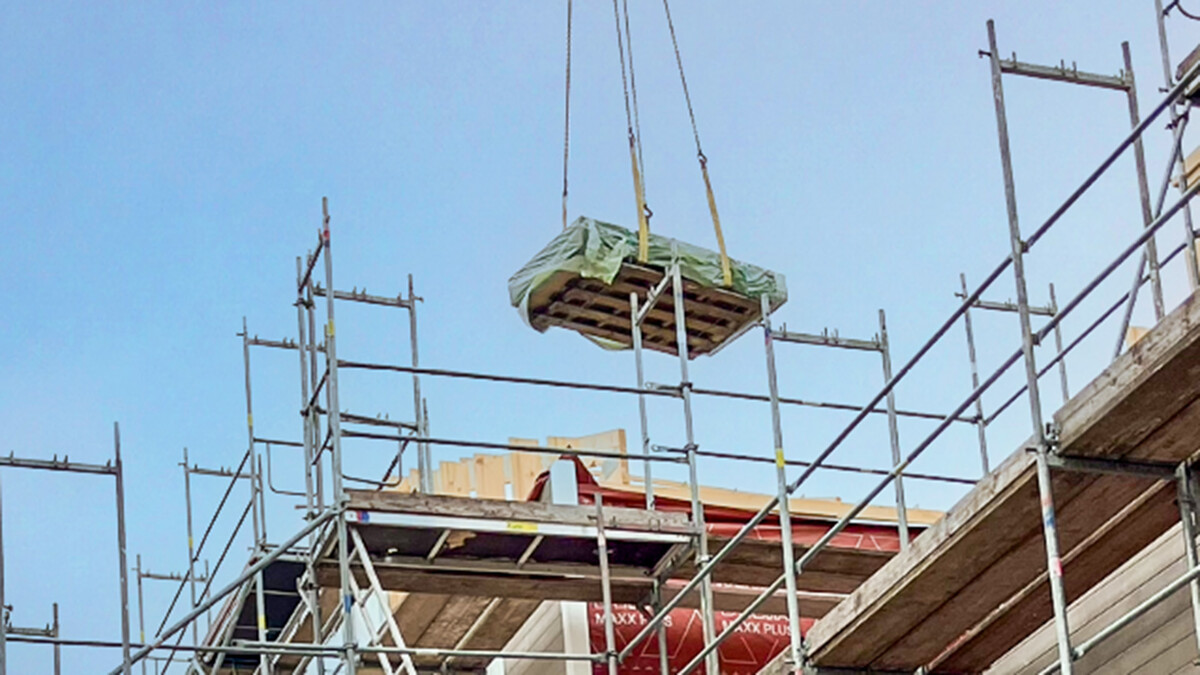 Der obere Bereich einer Baustelle, auf der ein hölzernes Gebäude errichtet wird. Man sieht Holzwände und Metallgerüste. Vor dem blauen Himmel eine Palette voller Baumaterialien, mit einer Plane abgedeckt und an einem Kran hängend - den Kran selbst sieht man allerdings nicht.
