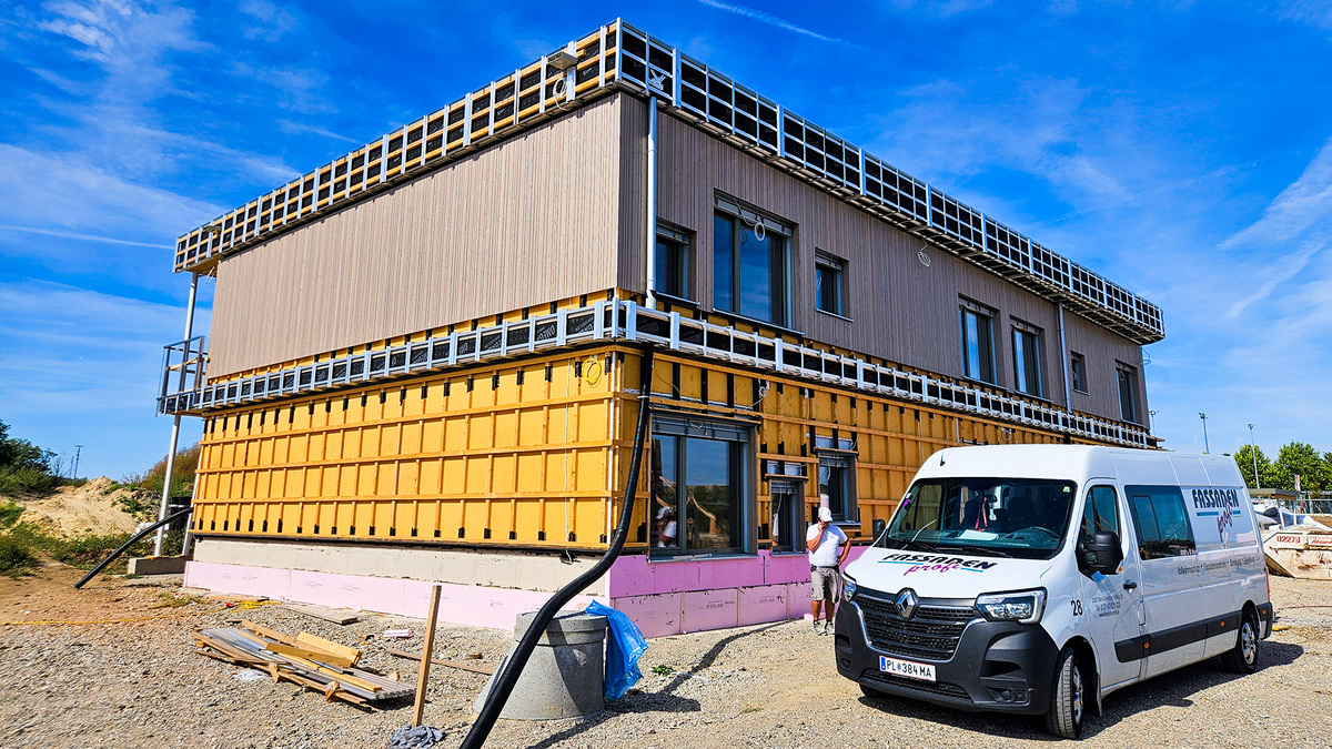 Ein neu gebautes zweistöckiges Bauwerk; in der Umgebung sind noch Bauwerkzeuge, ein Sprinter und der nackte Kiesboden mit Kabeln und Rohren. Das Gebäude selbst besteht größtenteils aus Holz; im unteren Stockwerk ist ein Gitterwerk aus Latten zu sehen, das obere ist bereits mit einer hölzernen Fassade ausgestattet. An einer Seite des Bauwerks sind außerdem Fenster und Türen zu sehen, einigermaßen unregelmäßig angeordnet.