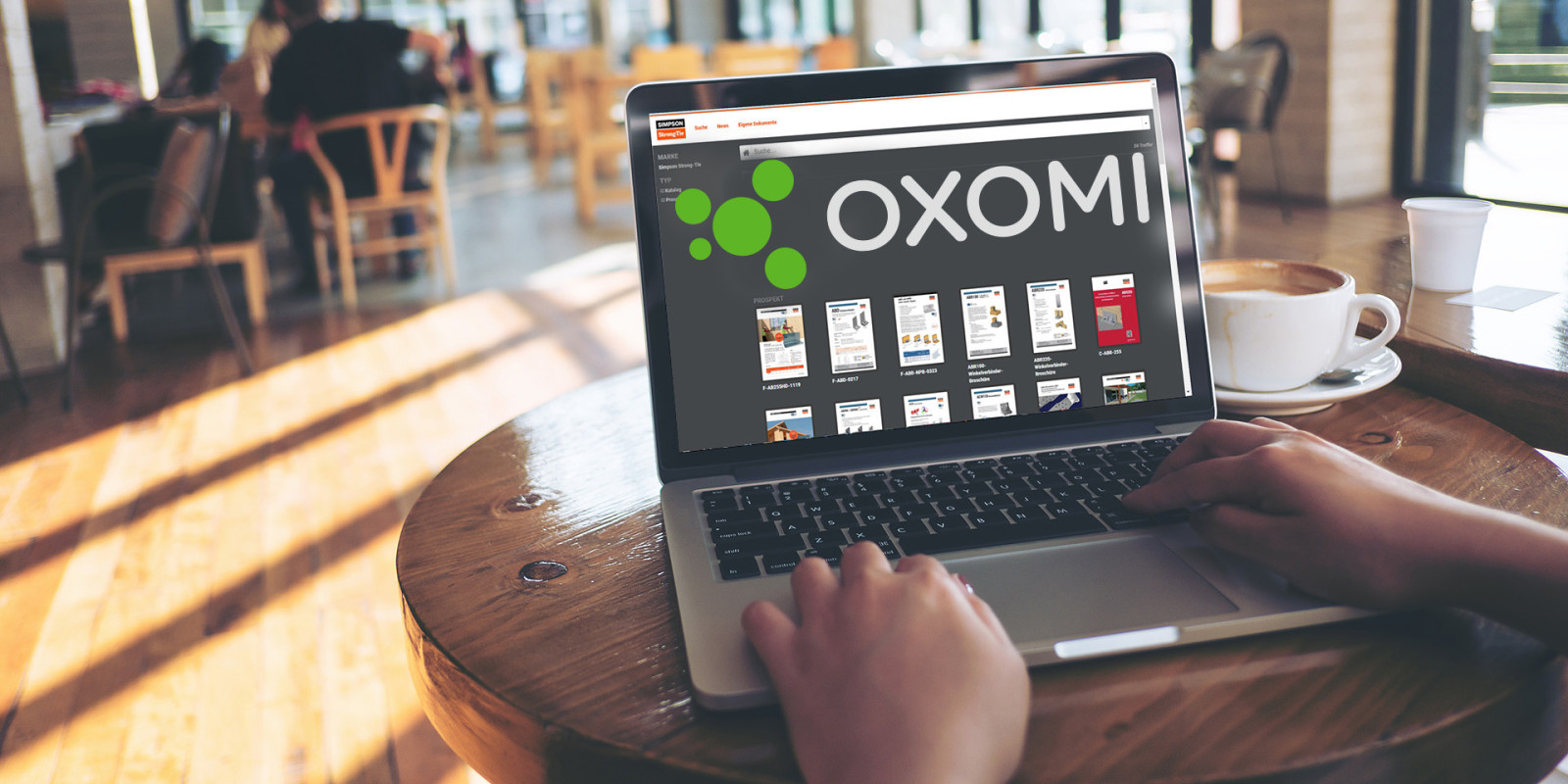 Ein Notebook auf einem Holztisch in einem gemütlichen Café. Auf dem Bildschirm des Notebooks sind das Oxomi-Logo sowie Produktinfos von Simpson Strong-Tie zu sehen.