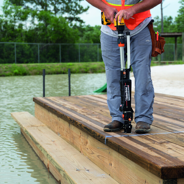 Ein hölzerner Steg oder Kai mit einer angerosteten Leiter am Ende, die in eine kleine Wasserfläche hinabführt. Auf dem Holz sieht man die Beine, ein Stück Unterkörper und die Arme einer Person in Arbeitsklamotten (unter anderem eine signal-orangene Sicherheitsweste). Die Person hält einen Timber Drive einsatzbereit, mit dem flachen Anschlag montiert. Ein Abschnitt des Kais ist mit blauer Kreide gekennzeichnet. Hintergrund: die kleine Wasserfläche, dahinter ein Stück Rasen, Maschendrahtzaun und Bäume.