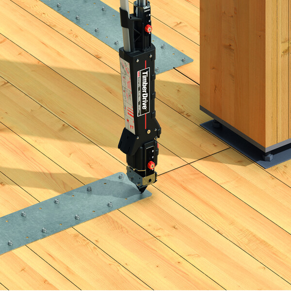 Eine große Holzfläche mit zwei Metallbändern, die jeweils von oben links und links unten kommen. Eine auf einem metallenen Abstandshalter stehende Holzsäule oder ein Pfeiler ist am rechten Bildrand sichtbar. Ein Timber Drive, fast komplett sichtbar, kommt von oben, um eine Schraube in das Metallband unten links einzudrehen. Die Präzisionsspitze ist sichtbar.