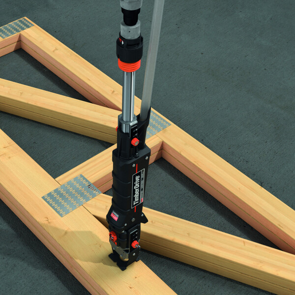 Eine Fachwerkkonstruktion aus Balken und Nagelplatten liegt auf einem dunklen, blaugrünen Boden. Ein Timber Drive steht aufrecht auf dem Holz, fast komplett sichtbar, mit dem flachen Anschlag vormontiert.