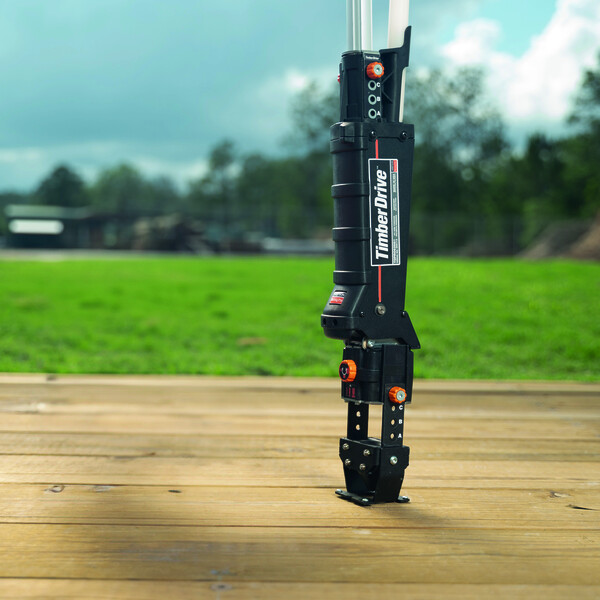 Der Timber Drive auf einer Holzfläche, ein Tisch oder eine Werkbank. Das Gerät steht eher rechts auf dem montierten flachen Anschlag und ist aufrecht, sodass nach oben nur das Laderohr und die Schrauber-Aufnahme sichtbar sind. Nach links hin ist der Tisch leer. Hintergrund, leicht verschwommen: eine grüne Wiese, Bäume, Büsche und Gebäude und darüber der blaue, leicht bewölkte Himmel.