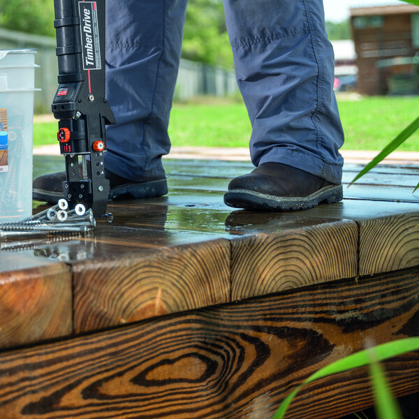 Ein Steg oder Dock aus Holz, mit einem Feuchtigkeitsfilm überzogen. In der rechten unteren Ecke die grünen Halme von Wasserpflanzen. Auf dem Holz sieht man die Beine einer Person in Arbeitsklamotten, die einen Timber Drive ans Holz ansetzt. Der Timber Drive ist mit dem flachen Anschlag ausgestattet, und einige Schrauben mit sechseckigen Löchern liegen bereits zum Verarbeiten bereit.