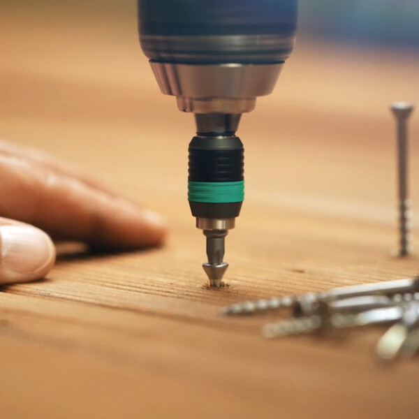 Der Kopf eines motorisierten Schraubers kommt von oben ins Bild und dreht eine Schraube in eine Holzfläche ein. Auf der rechten Seite sind weitere Schrauben zu sehen, eine davon mit der Spitze im Holz. Auf der linken Seite der Teil einer Hand, abgeschnitten vom Bildrand.