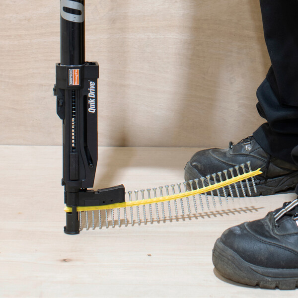 Ein Quik Drive-Vorsatz, der von oben ins Bild kommt und mit einem Streifen magazinierter Schrauben ausgestattet ist. Eine davon schraubt er in den hölzernen Boden ein. Auf der rechten Seite ist ein Stück der Beine des Anwenders zu sehen.