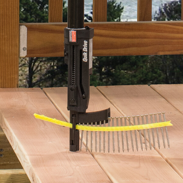 Ein Quik Drive-Vorsatz kommt von oben ins Bild, bestückt mit einem Streifen magazinierter Schrauben. Eine davon dreht er in eine Terrassendiele ein. Im Hintergrund ist ein hölzerner Zaun zu sehen, außerdem Bäume oder Büsche - das Ganze findet im Freien statt.