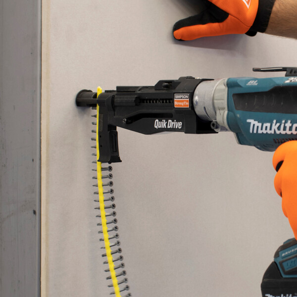 Ein blauer motorisierter Schrauber mit Quik Drive-Vorsatz, wiederum ausgestattet mit einem Streifen magazinierter Schrauben. Eine Schraube wird gerade in eine graue Faserzementplatte eingedreht. Rechts sind die Hände des Anwenders teilweise zu sehen; sie stecken in orangenen Handschuhen.