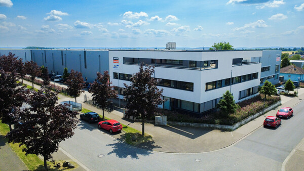 Ein von oben ausgeführter Schnappschuss des Firmengebäudes von Simpson Strong-Tie in Bad Nauheim. Das Gebäude ist von der vorderen Ecke aus sichtbar, hinten links beginnt der Lagerbereich. Im unteren Bereich eine Straße mit vereinzelten Autos und Bäumen.