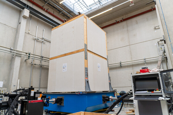 Ein simuliertes zweistöckiges Gebäude aus hölzernen Plattenelementen steht auf einer massiven Rüttelplatte, die blau lackiert ist. Das Gebäude ist quaderförmig, hat weder Fenster noch Türen. Der Hintergrund ist voller Geräte und Maschinen - ein Labor oder eine Werkstatt.