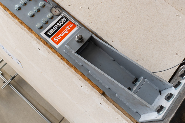 Ein massiver metallener Zuganker, der das Logo von Simpson Strong-Tie trägt. Er ist mit mehreren ZYK Schrägverschraubungen an der Holztafelwand dahinter befestigt, nur an einer Stelle fehlt die Schraube - dort sitzt stattdessen ein Messfühler mit einem Kabel.