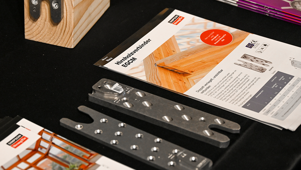 Auf einer mit einer schwarzen Tischdecke bedeckten Tischplatte liegen verschiedene Objekte: die beiden Hälften eines zweiteiligen Holzverbinders, ein Produktflyer zum Holzverbinder EGCM von Simpson Strong-Tie. Oben links ist ein hölzernes Modell zu erahnen, unten links eine aufgeschlagene Broschüre.