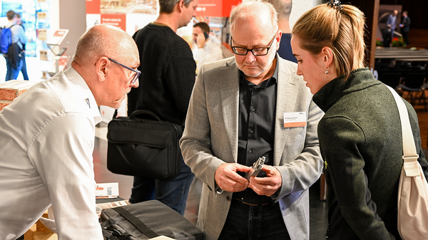 Das Bild zeigt drei Personen, die miteinander im Gespräch sind. Ein Mann hält einen zweiteiligen Holzverbinder und mustert ihn näher, ein zweiter Mann beugt sich von links heran und scheint das Teil zu erklären. Eine Frau beugt sich von rechts heran und mustert das Teil ebenfalls interessiert.