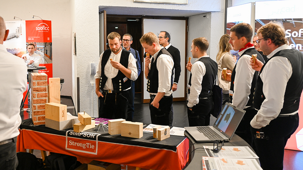Eine Gruppe von Personen in Zimmererkluft kommt von rechts ins Bild, jeder hält eine Eistüte in der Hand. Auf den linken zwei Drittel des Bildes ist ein Tisch vom Messestand von Simpson Strong-Tie mit einer schwarz-orangeroten Tischdecke zu sehen, darauf jede Menge Broschüren, Produkte und Modelle. Die Zimmerer ganz links in der Reihe schauen nicht mehr auf ihr Eis, sondern auf das, was sich alles auf dem Tisch befindet. Hinter ihnen ist der Ausgang aus dem Raum zu sehen.