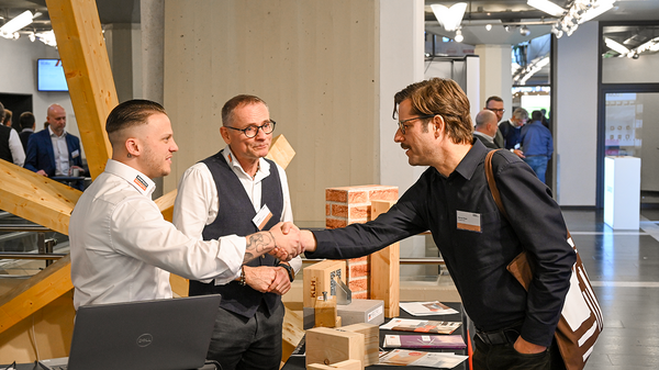 Das Bild zeigt drei Männer, von denen sich zwei über die Messetheke von Simpson Strong-Tie hinweg die Hand geben. Ein dritter Mann befindet sich ungefähr zwischen den beiden und lächelt zufrieden. Hinter dem Mann ganz links ist ein hölzernes Modell mit vielen von einem zentralen Element ausstrahlenden hölzernen Balken.