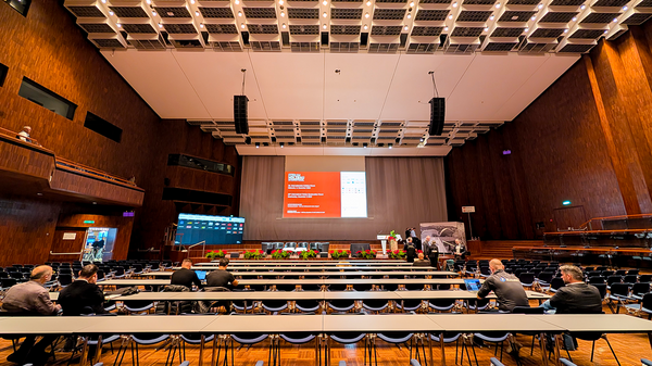 Die Bühne in einem Vortragssaal, davor zahlreiche Reihen Tische und Stühle. Vorne auf der Bühne eine Leinwand mit einer roten Projektion; deutlich zu sehen die Beschriftung "Forum Holzbau International". Die Wände des Raumes sind aus dunklem Holz.