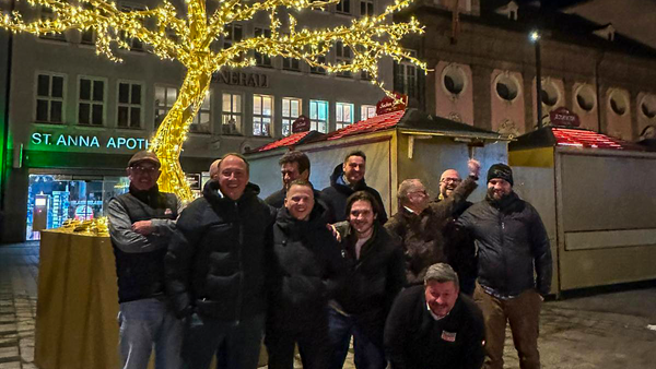 Eine Gruppe Menschen in warmen Klamotten stehen oder hocken vor einem mit weihnachtlichen Lichtern geschmückten Baum. Hinter ihnen sind Hütten eines Weihnachtsmarktes zu sehen. Das ganze Bild ist eher dunkel; es scheint Abend oder Nacht zu sein. Die Personen n der Gruppe lachen fröhlich.