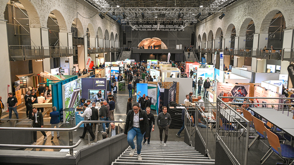 Eine Messehalle voller farbenfroher Stände und Banner, von schräg oben betrachtet. Eine Treppe führt vom Betrachter hinunter zu den Ständen. Viele Leute sind unterwegs. Links und rechts tragen große Steinbögen die Decke; es scheint sich um ein historisches Gebäude zu handeln. 