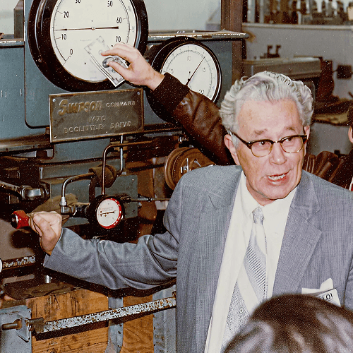 Produktleiter Tye Gilb auf einem Foto aus den Sechzigern. Er trägt ein graues Jackett und steht vor einer Maschine voller Skalen und Rohre.