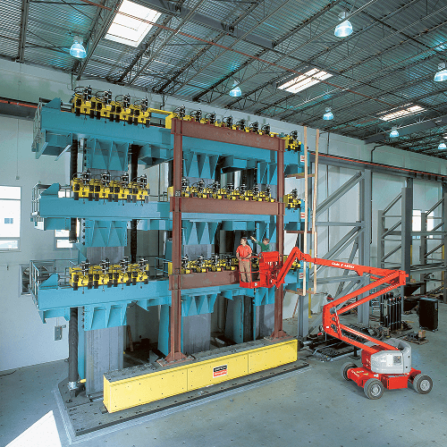 Ein Labor, gefüllt mit einem regalartigen Aufbau aus massiven, blau lackierten Stahlelementen. Eine Reihe gelber Geräte, möglicherweise Messsysteme, ist daran befestigt. Zwei Personen in einem Hebelift befinden sich auf halber Höhe des Aufbaus , einer der beiden zeigt darauf.