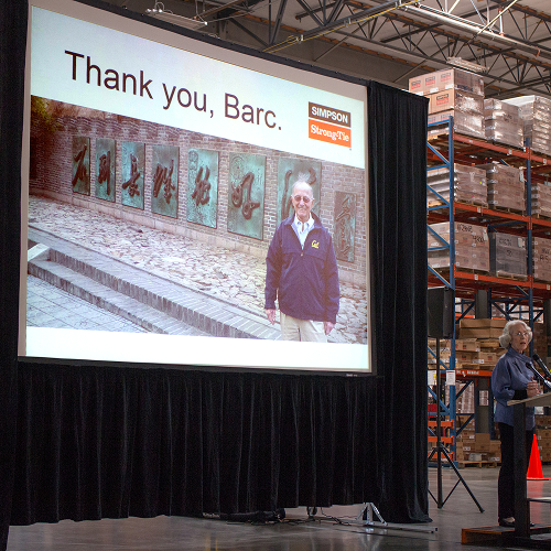 Das Bild zeigt das Innere eines Lagers - rechts sind gefüllte Hochregale zu sehen. Fokus ist jedoch eine Leinwand, auf die ein Bild von Barclay Simpson projiziert wird, zusammen mit der Aufschrift "Thank you, Barc." sowie dem Logo von Simpson Strong-Tie.