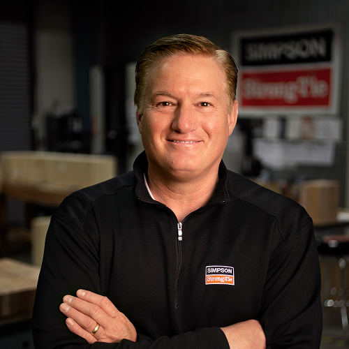 Präsident Mike Olosky mit verschränkten Armen und einem schwarzen Hemd oder Sweatshirt, auf dem das Simpson Strong-Tie Logo zu sehen ist. Er lächelt in die Kamera.