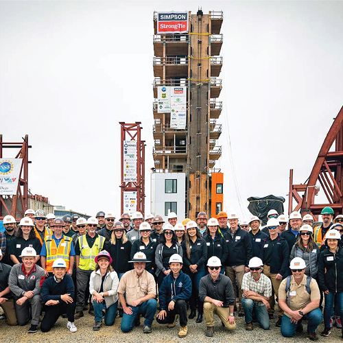 Eine große Gruppe Menschen, alle mit Bauhelmen bekleidet, steht und kniet, Richtung Kamera blickend, vor einem mehrstöckigen, offenen Holzturm, an dem ganz oben eine Fahne mit dem Simpson Strong-Tie-Logo hängt.