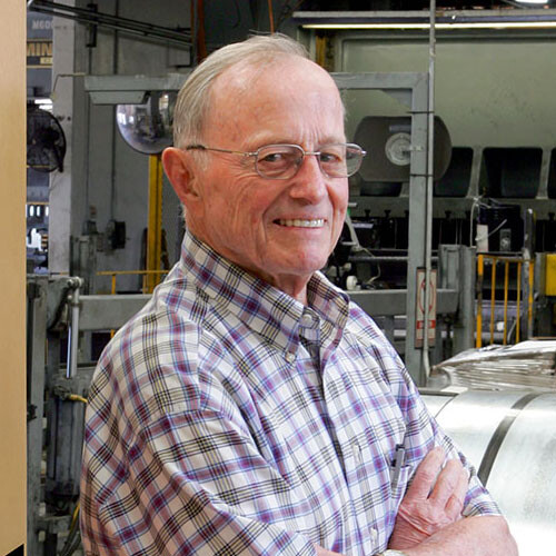 Der Gründer von Simpson Strong-Tie, Barclay Simpson, in einem karierten Hemd und mit verschränkten Armen. Er lächelt in die Kamera. Im Hintergrund ist eine Werkstatt oder Produktionsstätte zu sehen.