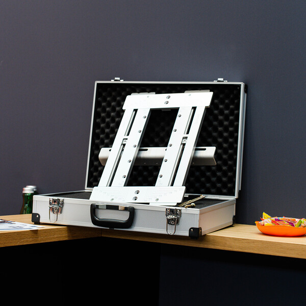 In einem aufgeklappten Metallkoffer, ausgepolstert mit schwarzem Schaumstoff, liegt ein Werkzeug oder Hilfsmittel aus glänzendem Aluminium, geformt wie ein Rechteck. Daneben eine kleine Schüssel Süßigkeiten.