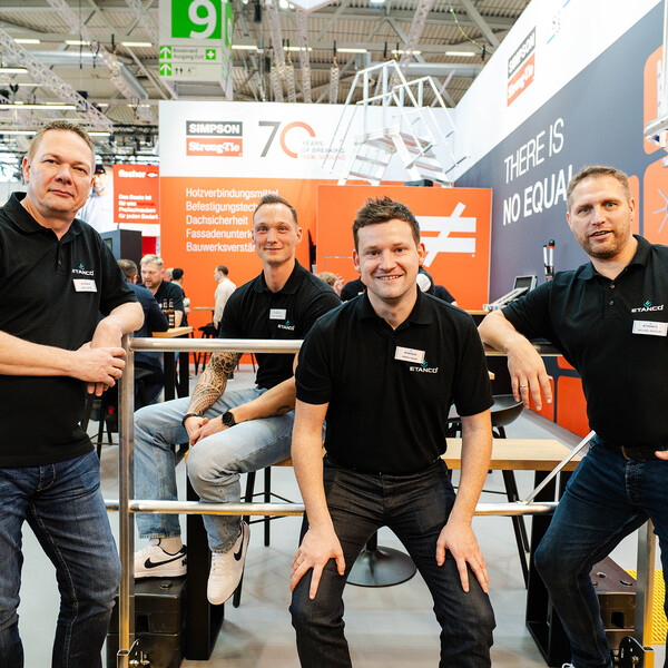 Vier Personen in schwarzen Polos und in Jeans stehen bzw. sitzen nebeneinander und lächeln in die Kamera. Auf den Hemden ist das Logo der Firma Etanco zu sehen. Im Hintergrund der Trubel einer großen Fachmesse.