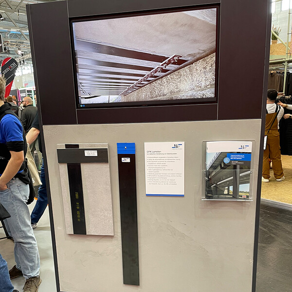 Eine Produktwand mit Carbon-Lamellen und einem Modell, das die Anwendung einer solchen Lamelle demonstriert. Darüber ein Bildschirm. Im Hintergrund sich umherbewegende Menschen.