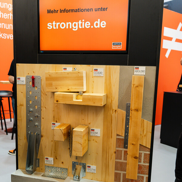 Eine Produktwand mit einer Oberfläche aus Holz, an der diverse Holzverbinder befestigt sind, zusammen mit den dazugehörigen hölzernen Bauelementen. Ein Teil der Wand stellt Beton oder Ziegelwerk dar. Oberhalb des ganzen ein Bildschirm, der den Text "Mehr Informationen unter Strongtie.de" zeigt. Die Verbinder sind: Universalverbinder CCS, Hirnholzverbinder EGCL & EGCM, Sanierungswinkel ABGU360, Zuganker HDCLT140, Winkelverbinder ABCLT210 und Zuganker HTT140.