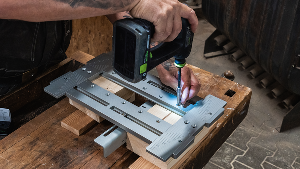 Zu sehen: eine Schablone aus Metall (Aluminium) auf einer hölzernen Werkbank. Zwei tätowierte Arme kommen von links ins Bild, einen elektrischen Bauschrauber und eine Schraube haltend. Sie sind damit beschäftigt, die Schablone auf einem Bauelement aus Holz zu befestigen. Bei der Schablone handelt es sich um die MIMO315 von Simpson Strong-Tie.