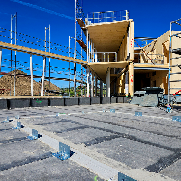 Der Betonboden einer Baustelle; offenbar entsteht hier ein Innenraum. Auf dem Beton sind bereits mehrere Winkelverbinder befestigt, die zum Anschluss von Holzwänden dienen. Im Hintergrund sind Baugerüste sowie einige bereits konstruierte Räume zu sehen.