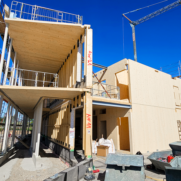 Eine Holzbaustelle in zwei Stockwerken, die auf einem Fundament aus Beton ruhen. Wände, Decken und Stützkonstruktion bestehen komplett aus Holz. Im Hintergrund ist ein Kran zu sehen, im Vordergrund Container.