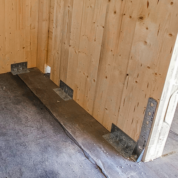 Eine Holzwand, die rechts endet - vermutlich an der Türöffnung. Links ist die Ecke des Raumes zu sehen. Mehrere Verbinder aus dunkelgrauem Metall verankern die Holzwand mit dem Boden aus Beton.