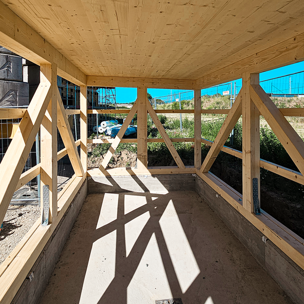 Eine Holzbaustelle bzw. ein einzelner Raum in dieser Baustelle. Die Wände sind aus Fachwerk-ähnlichen Holzbalken, die Decke besteht komplett aus Holz. Der Boden und die einige Zentimeter hohe Aufkantung ist aus Beton. Außerhalb des Raums zu sehen: grüne Natur und ein blauer Himmel.
