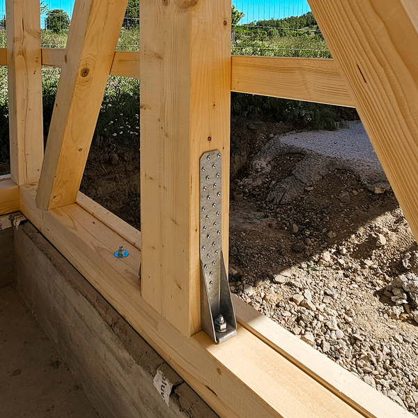 Eine Fachwerkwand aus Holzbalken auf einer Aufkantung aus Beton. Ein einzelner Zuganker hält einen aufrecht stehenden Balken mit dem darunter befindlichen Querbalken verbunden. Im Hintergrund sind grüne Natur und ein blauer Himmel zu sehen.