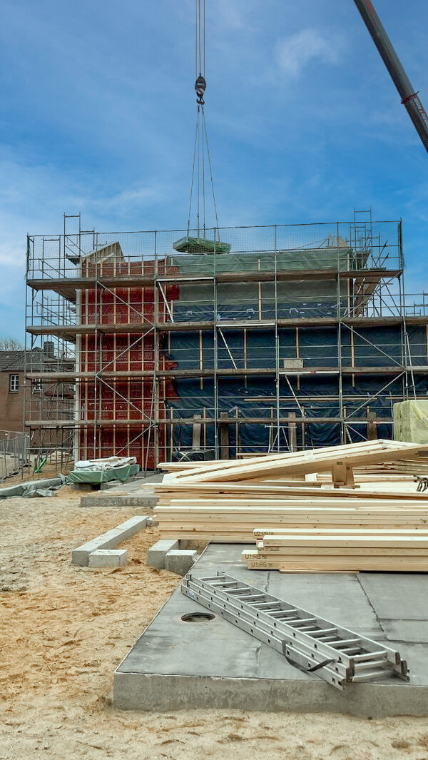 Eine Baustelle, auf der ein Holzhaus entsteht. Im Hintergrund das Gebäude selbst, umgeben von Gerüsten und von einem Baukran bedient. Im Vordergrund ein Betonfundament, darauf liegend eine zugeklappte Stehleiter aus Aluminium sowie mehrere Stapel Holzbalken und Holzbauteile.