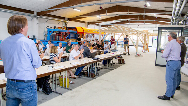 Das Innere einer Fabrik- oder Werkshalle. An langen Tischreihen sitzen zahlreiche Menschen mit Namensschildern und applaudieren. Im Hintergrund ist eine Konstruktion aus schlanken, hellen Holzbalken zu sehen, wie das Skelett eines kleinen Hauses. Dahinter, an der Wand des Raumes, eine Abbundanlage und weitere Maschinen.