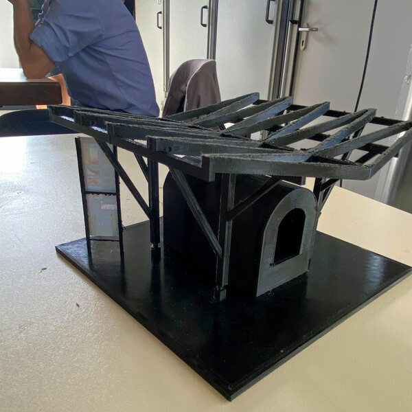 Auf einer Tischplatte steht ein aus schwarzem Plastik gedrucktes 3D-Modell eines Bauwerks - eine Röhre (oder so ähnlich) mit einem eckigen Boden und rundem Oberteil wird von einem Dach auf vier Stützen getragen. Im Hintergrund sind metallene Schränke sowie eine sitzende Person in einem blauen T-Shirt.