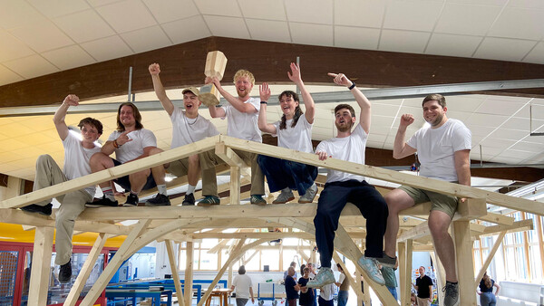 Auf einer Holzkonstruktion sitzt eine große Gruppe Personen, Männer und Frauen, und jubelt. Der in der Mitte - auf dem Giebel - sitzende hält eine Art Pokal aus Holz hoch. Im Hintergrund ist zu sehen, dass die ganze Szenerie sich im Inneren eine Werk- oder Fabrikhalle abspielt. Im Hintergrund stehen außerdem noch weitere Personen.